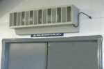 A wall-mounted air conditioning unit is located above a gray door, next to a Vestil Variable Speed Air Curtain 48 In Width. A blue sign reading Excellence is partially visible below, on the crisp white wall.