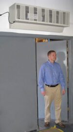 A man in a blue shirt and beige pants stands in a doorway beneath a wall-mounted air conditioning unit, featuring the efficient Vestil Variable Speed Air Curtain 48 In Width. The room is framed by gray walls and doors.