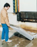 A person in a brown jacket and blue jeans holds a metal ramp near a loading dock featuring a Vestil Wheel Riser Optional Portable Wheel. Snow and slush cover the ground around the dock, which has a black bumper. The person seems ready to position the ramp.