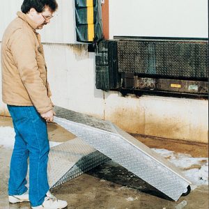 A person in a brown jacket and blue jeans holds a metal ramp near a loading dock featuring a Vestil Wheel Riser Optional Portable Wheel. Snow and slush cover the ground around the dock, which has a black bumper. The person seems ready to position the ramp.