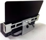 A laptop docked in a metallic horizontal docking station, featuring adjustable sliding grips and a central locking mechanism, placed on a light-colored surface.