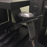 Handheld barcode scanner resting in a black holder attached to a vertical rack with multiple power outlets. The scanner is positioned on a shelf amid electronic equipment.