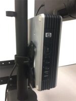 A compact HP desktop computer is mounted vertically on a stand, positioned behind a monitor. The front panel shows ports, a power button, and the HP logo. The stand is attached to a base on a flat surface.