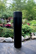 A Vestil Cinco-Black Bollard Cover 52 In stands on a paved surface with stone-bordered landscaping. Lush greenery and colorful flowers are abundant in the background under a canopy of trees.