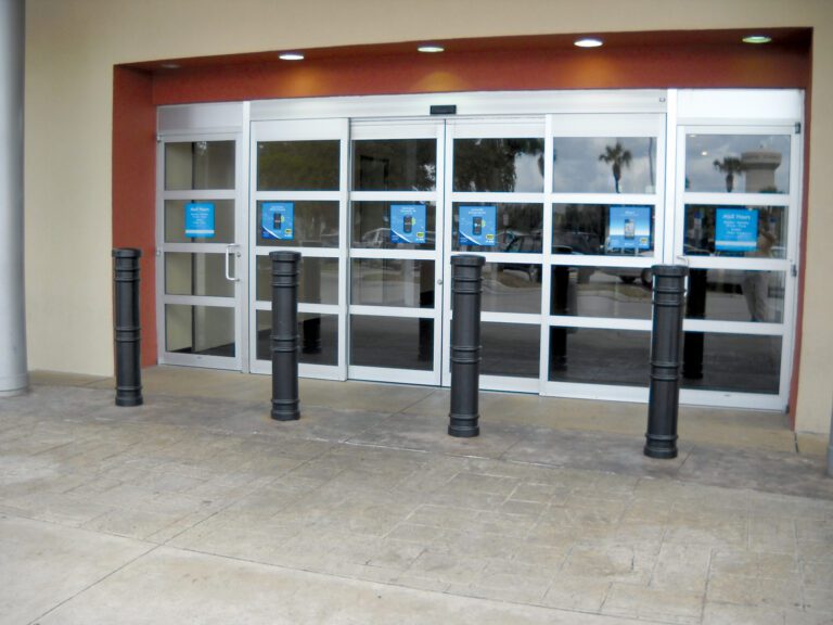 black deco bollard outside store entrance Automatic sliding glass doors with reflective panels are set against a soft yellow and red wall, protected by five black bollards each with a 52-inch Vestil Metro-Black Bollard Cover, on patterned stone pavement. Blue-background signs adorn each door panel.