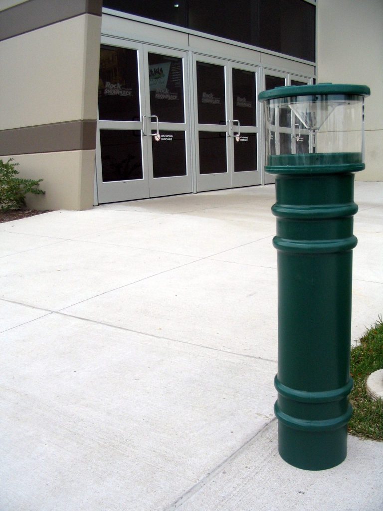 BPC-DM-LAC-FG_A.jpg A Vestil Metro-Green Bollard Cover-Ac Light 57 In, featuring a transparent section, stands on a concrete sidewalk near glass-door building entrance, with small plants visible in the background.
