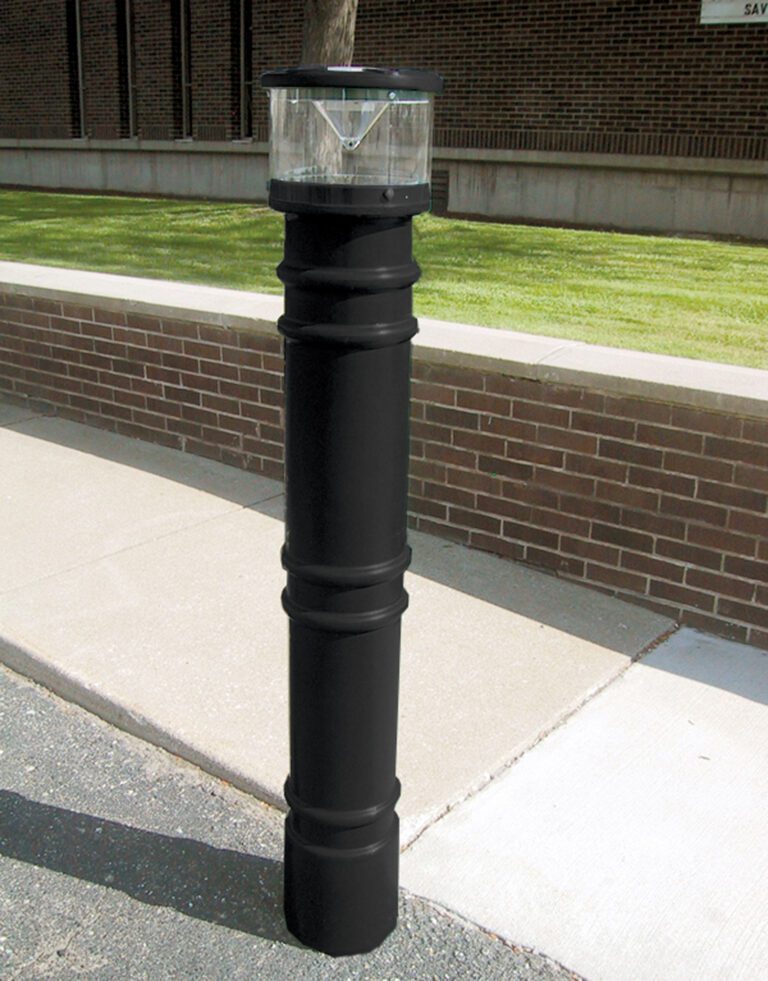 A paved pathway beside a brick wall features a black bollard with a transparent, dome-shaped light resembling the 57-inch Vestil Metro-Black Bollard Cover W/Light. Grass and a building can be seen in the background.