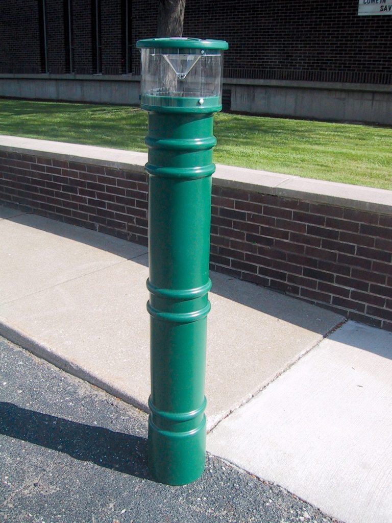 BPC-DM-LUV-FG_A.jpg A tall, green cigarette disposal bin stands on a sidewalk next to a brick wall. The design resembles a Vestil Metro-Green Bollard Cover W/Light 57 In, with a clear section at the top for viewing contents. Pavement and grass are visible in the background.