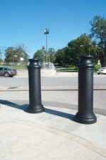 Two large, black cylindrical bollards on a sidewalk by a road with cars in the background. Trees, grass, and fencing under a clear blue sky complete the scene, enhanced by the sleek Vestil Pawn-Black Bollard Cover 49 In.