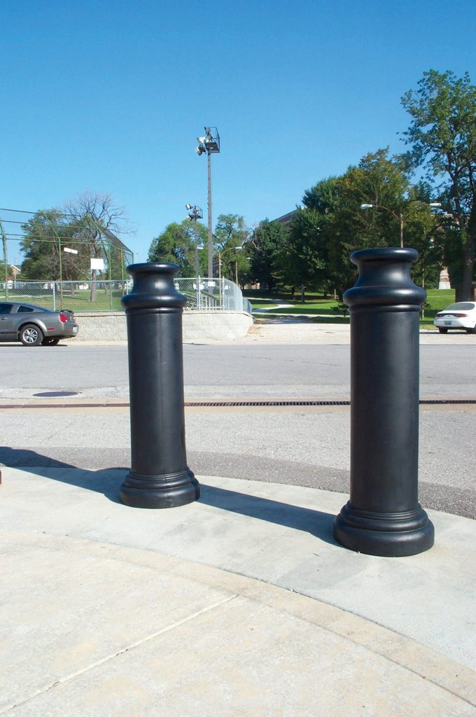 Two large, black cylindrical bollards on a sidewalk by a road with cars in the background. Trees, grass, and fencing under a clear blue sky complete the scene, enhanced by the sleek Vestil Pawn-Black Bollard Cover 49 In.