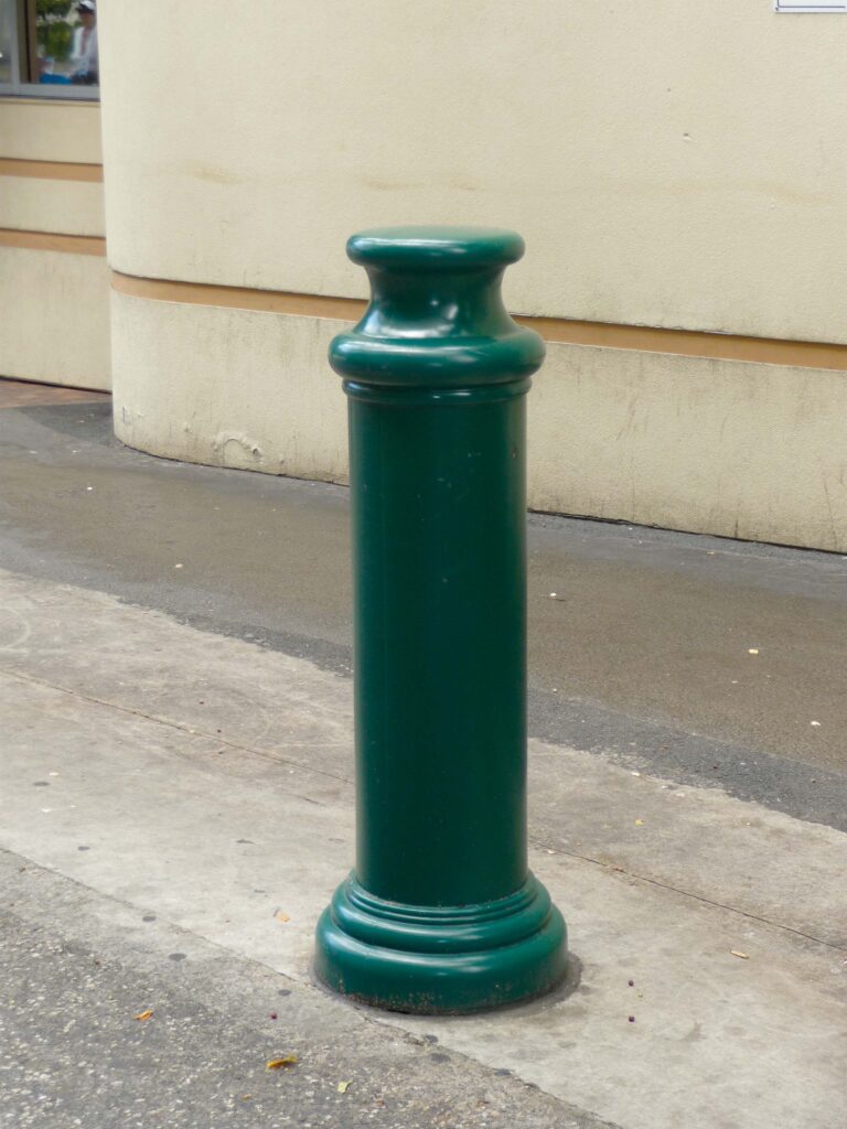 A Vestil Pawn-Green Bollard Cover 49 In stands on a concrete surface near a building, its tall, green cylindrical shape with rounded top enhancing modernity and safety along the sidewalk against the smooth wall backdrop.