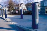 A row of gray bollards, featuring the Vestil Rib-Gray Bollard Cover 52 In, lines a paved area beside a street. Residential houses and trees form the backdrop, alongside a chain-link fence. The scene unfolds on a clear, sunny day.