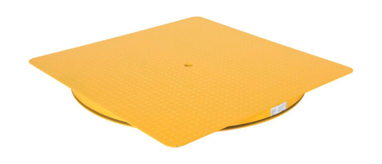 A large, yellow plastic lid with a textured surface and central hole rests on the ground, similar to a Vestil Hd King Pin Carousel. Its square top curves downwards with partially visible sides and a small label on one side.