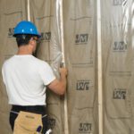 A person in a blue hard hat and white shirt is installing insulation in a wooden frame wall. They have a tool in hand, tool belt around their waist, next to bundled MH-USA 20 x 100 - 6 Mil Clear Poly Sheeting. The insulation bears the JM logo.