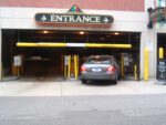A car enters a parking garage with two yellow clearance bars, including a Vestil Clearance Bar-No Lettering 78 In marked CLEARANCE 88. Upward arrows adorn the entrance sign, a parking fee sign decorates the right wall, and the area is bordered by a brick wall.