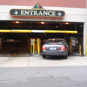 A car enters a parking garage with two yellow clearance bars, including a Vestil Clearance Bar-No Lettering 78 In marked CLEARANCE 88. Upward arrows adorn the entrance sign, a parking fee sign decorates the right wall, and the area is bordered by a brick wall.