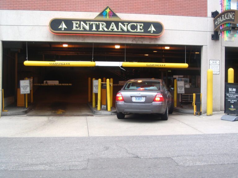 CLB-5-78P_A.jpg A car enters a parking garage with two yellow clearance bars, including a Vestil Clearance Bar-No Lettering 78 In marked CLEARANCE 88. Upward arrows adorn the entrance sign, a parking fee sign decorates the right wall, and the area is bordered by a brick wall.