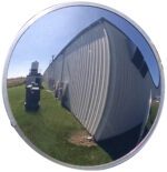 A Vestil Convex Mirror Outdoor 26 Ft Distance shows a warped view of a light industrial building, small gear, and nearby grass. The bright sky suggests sunshine, with visibility reaching up to 26 feet.