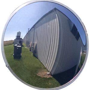 A Vestil Convex Mirror Outdoor 26 Ft Distance shows a warped view of a light industrial building, small gear, and nearby grass. The bright sky suggests sunshine, with visibility reaching up to 26 feet.