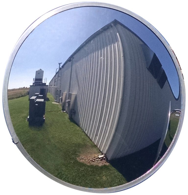 A Vestil Convex Mirror Outdoor 26 Ft Distance shows a warped view of a light industrial building, small gear, and nearby grass. The bright sky suggests sunshine, with visibility reaching up to 26 feet.