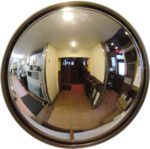 A fisheye view of a narrow, dimly lit corridor shows a Vestil Convex Mirror Outdoor 16-20 Ft 4 In Deep. It reflects the kitchen on the left and the seating area on the right, distorting kitchen equipment and chairs along this elongated passage.