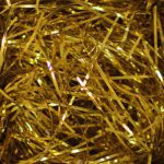A close-up of tangled 10 lb. Gold Metallic PureMetallic Shred™ (10 lbs/case) strands forms a shiny, reflective surface, with an intricate web-like pattern that captures light and highlights the metallic texture and bright yellow hues.