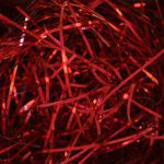 The 10 lb. Red Metallic PureMetallic Shred™ Veryfine Cut™ - 10 LBS PER CASE features a shiny array of thin red strands, akin to festive tinsel, that brilliantly reflect light and create a vibrant shimmer against a dark background.