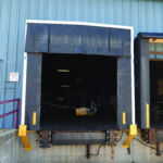The industrial loading dock includes a raised bay door with a black protective curtain. It features a blue structure with yellow bumpers and a chain barrier. Securing the area efficiently is the Vestil Dock Shelter 24 In Projection 11X10.5 Ft.