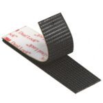 A close-up of a 1 x 150 Black 3M™ SJ3540 Dual Lock™ Fastener (2 Rolls/Case) shows its textured surface with one side curled up and the white adhesive backing with red text underneath. This fastener offers strong adhesion, enhancing industrial applications.