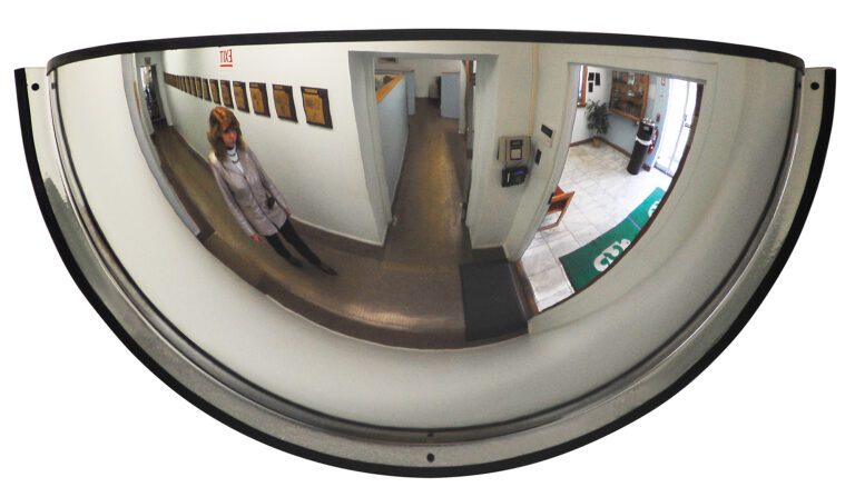 A woman is reflected in a Vestil Dome Mirror 180 Degree 32 Ft Distance while walking down a hallway with framed pictures. An open area with chairs and a rug is visible in the reflection.