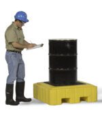 A person in a blue hard hat, safety goggles, tan shirt, jeans, and black boots writes on a clipboard next to a black barrel on a yellow Vestil Ultra Spill Pallet.