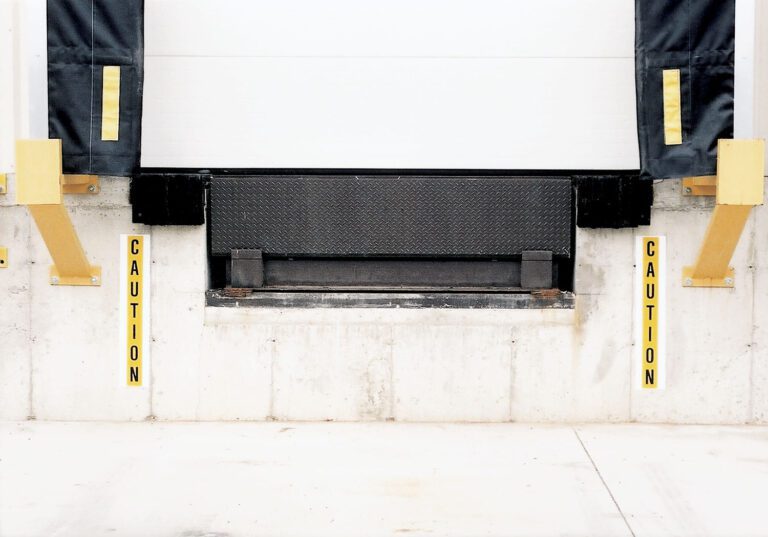 ÊÊÉÉ The loading dock entrance has a closed white door, flanked by two yellow Caution signs on the pillars. Black padding frames the door for added safety against the concrete ground, while 28-inch high Vestil Dock Safety Reflective Stripes ensure visibility nearby.