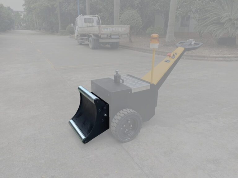 A road cleaning machine with a black scoop and large wheels, featuring a yellow handle and small control panel similar to the Vestil Roll Pusher (16 width), is parked on concrete. A truck framed by roadside trees is visible in the background.