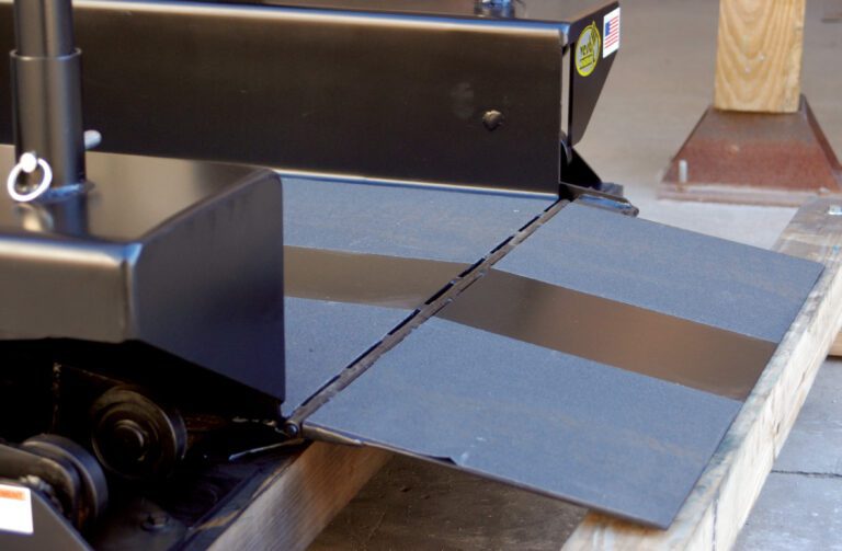 A close-up showcases a black metal loading ramp, like the Vestil Optional Steel Rollover 52 X 12, secured to a machinery platform. The lowered ramp has a textured surface for traction, with part of a wooden post and cement floor seen in the background.