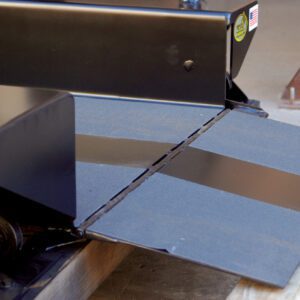 A close-up showcases a black metal loading ramp, like the Vestil Optional Steel Rollover 52 X 12, secured to a machinery platform. The lowered ramp has a textured surface for traction, with part of a wooden post and cement floor seen in the background.