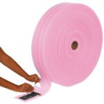 A person wraps a small electronic component with an MH-USA 1/8 x 6 x 550 (12) Perforated Anti-Static Air Foam Roll. The pink foam, partly unrolled, shows its thickness and texture as the component rests securely at the tables edge.