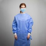 A person with red hair stands facing forward, wearing a blue medical gown and a blue face mask. The background is a plain, light-colored wall.