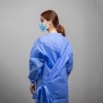 A person with long hair tied back wears a blue medical gown and a face mask, viewed from the side. The gown is tied at the back. The background is neutral gray.
