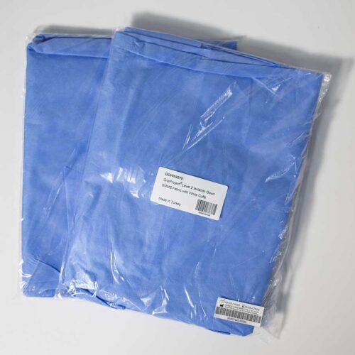 Two packages of folded blue scrub tops are sealed in transparent plastic. Each package has a white label displaying text. The fabric appears smooth and is neatly packed, with the tops intended for medical or professional use.