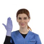 A person with reddish-brown hair wearing blue medical scrubs and a purple glove on one hand is smiling and holding up the gloved hand. The scrubs have a logo that reads GripProtect. The background is plain white.