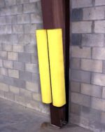 A steel pole, guarded by two yellow Vestil Polyethylene Ultra-I-Beam Protectors, stands in a corner against a brick wall and smooth concrete floor, ensuring safety and durability.