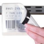 A hand peels back a clear cover from a label holder on a shelf, revealing numbers, a barcode, and 8465-2497D, subtly hinting at MH-USAs versatile 8 1/2 x 11 magnetic tape strips for seamless organization.