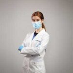 A woman in a white lab coat and blue medical mask stands with arms crossed against a plain background. She wears blue gloves and has auburn hair tied back.