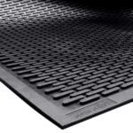 A close-up of the black MH-USA 3 x 5 Scraper Entry Mat shows raised horizontal lines with a textured design. The edge is slightly lifted, revealing its smooth underside.