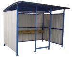 The compact metallic bus shelter echoes the design of the Vestil Multi-Duty Smokers Shelter 120 In, with a curved roof, blue frame, and white panels. It features an open front and a wooden bench for comfort against the back wall while standing on sturdy support legs.