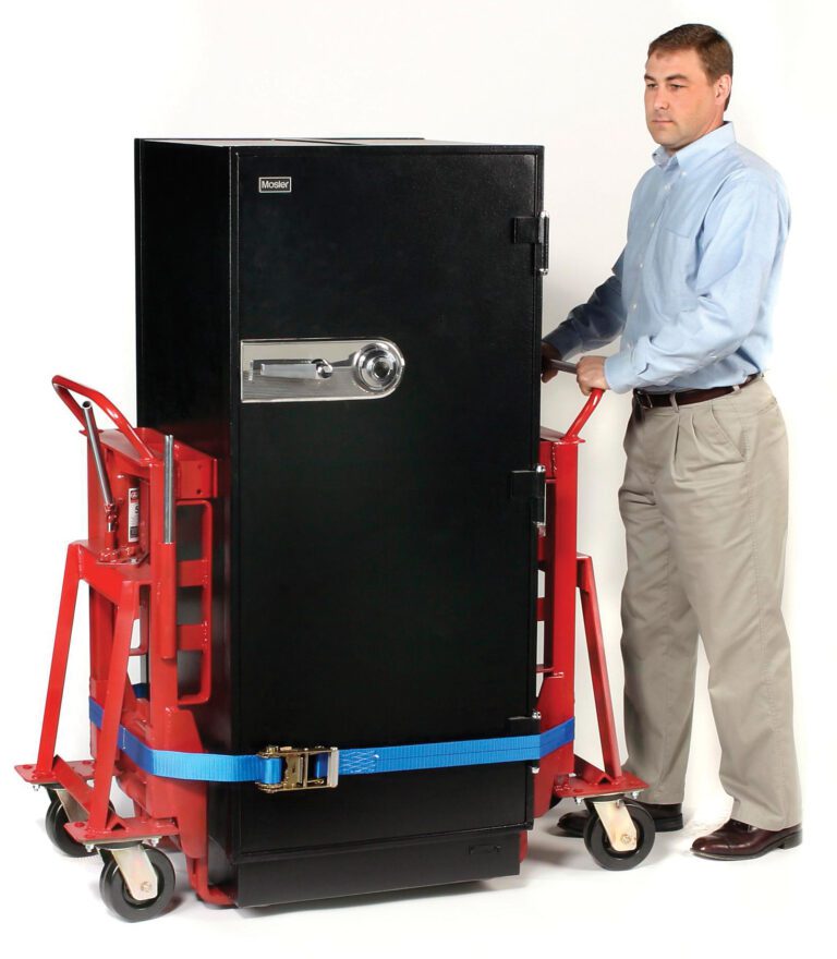 A man in a light blue shirt and beige pants uses a red Vestil Vend. Machine-Furn. Movers 6000 Lb Cap hand truck to move a large black safe, secured with blue straps, against a plain white background.