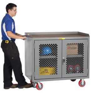 A person in a blue shirt and black pants, with a tape measure at their belt, pushes a Little Giant Mobile Bench Cabinet with Clearview Doors. The gray cabinet, loaded with tools and equipment, moves smoothly on red casters as they navigate the space.