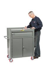 An older adult in glasses, a blue shirt, and jeans writes on a Little Giant Mobile Bench Cabinet with Heavy-Duty Drawer. The gray cabinet has red wheels, a drawer, two doors, and a side handle against a white backdrop.