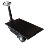 The Vestil Traction Drive Cart Platform 0.75K Lb features a black flatbed design, two large wheels, and a steering handle with intuitive controls. The platform has a diamond plate texture, and the handle is attached to one end of the cart.