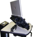 A laptop is mounted on a mechanical arm positioned at an angle on a desk. The setup is designed for ergonomic adjustment. A small speaker or electronic device is visible on the desk to the left.